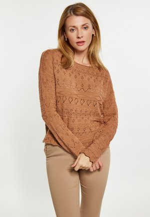 Gebreide trui in warm bruin met een gestructureerd patroon, voorzien van opengewerkte ontwerpen, lange mouwen en een ronde halslijn, gecombineerd met beige broeken.