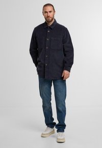 Mann steht da in einem dunklen, geknöpften Cordhemd, blauen Jeans und weißen Sneakers, vor einem schlichten hellen Hintergrund.
