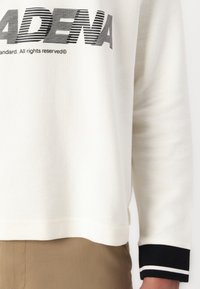 Bjela teksturirana dukserica s crnim prugastim detaljima na rukavima i izrazitim crnim natpisom "PASADENA" preko prsa.