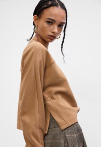 Tânăr cu păr împletit, purtând cercuri de aur, un hanorac mare de culoare maro și pantaloni în carouri, împotriva unui fundal simplu.