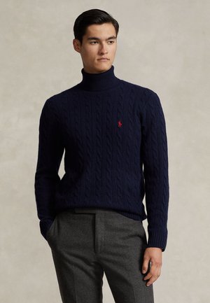 Homme portant un pull à col roulé bleu marine en maille torsadée et un pantalon gris, debout devant un fond beige uni.