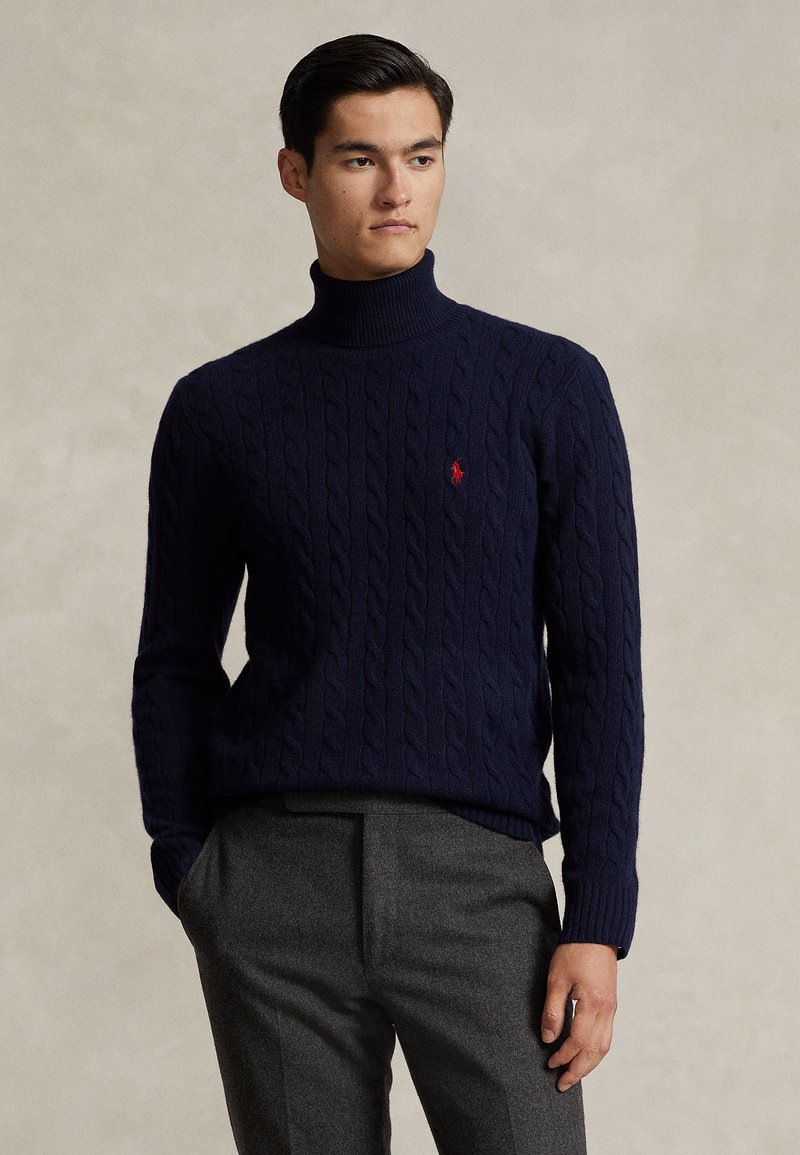 Uomo che indossa un dolcevita blu navy a trecce e pantaloni grigi, in piedi davanti a uno sfondo beige uniforme.