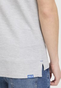 T-shirt gris clair en tissu doux, présentant une petite étiquette bleue "ISOLID" et des fentes sur les côtés à l'ourlet pour plus de détails.