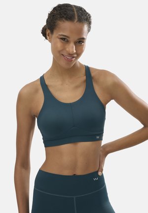 Mujer sonriente con sujetador deportivo de color verde azulado oscuro y leggings a juego, posando con una mano en la cadera sobre un fondo liso.