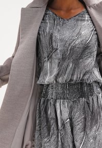 Šedý oversize kabát přes vzorovaný černobílý top. Top má výstřih do V a nařasený pas, což zdůrazňuje lehký design látky.