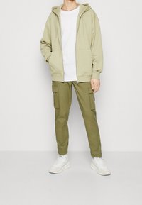 Sudadera verde oliva con cremallera y cordones, combinada con pantalones cargo a juego. Camiseta blanca por debajo, complementada con zapatillas blancas.