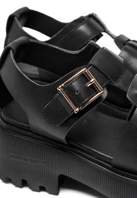 Black platform sandals with a wide strap design, featuring a metallic buckle accent and textured rubber soles with ridges.