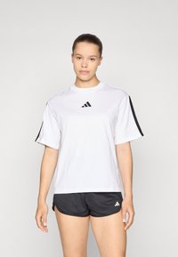 Bijela majica kratkih rukava sportske izvedbe s crnim detaljima i Adidas logom na prsima, u kombinaciji s crnim šortsevima s detaljem logotipa.