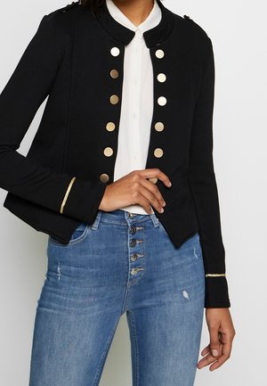 Kvinde iført sort militærstiljakke med guldknapper og kant, hvid bluse og højtaljede blå jeans med flere knapper.