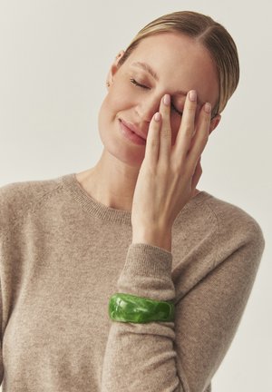 Grøn marmoreret armring lavet af glat resin båret på en nøgenfarvet sweater med ribbet kant. Teksturen er blank og tilføjer et markant accent.