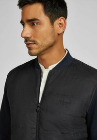 Dunkle gesteppte Bomberjacke mit kontrastierendem marineblauen Kragen, ausgestattet mit einem Frontreißverschluss und einem dezenten Logo auf der Brust, getragen über einem weißen Hemd.