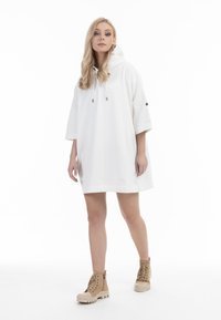 Rochie-hoodie albă supradimensionată cu mâneci scurte, ce dispune de o glugă, accente cu șnur și un detaliu cu logo pe partea laterală. Asortată cu cizme bej cu șireturi.