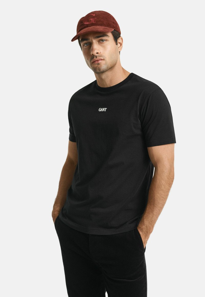 Man in een zwart GANT T-shirt, donkere broek en een rode pet, staande met zijn handen in de zakken tegen een effen witte achtergrond.