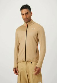 Bež jakna na zip s teksturiranom tkaninom, visokim ovratnikom i crnim patentnim zatvaračem. Sadrži narančaste akcentne detalje i logotip na prsima.