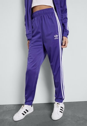 Persoon draagt paarse Adidas trainingsbroek met witte strepen en witte Adidas sneakers, staande tegen een grijze muur en vloer.