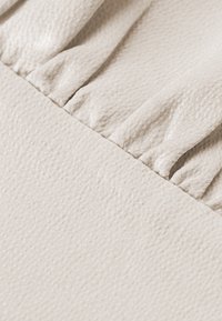 Tessuto beige chiaro con una superficie texturizzata. La parte superiore presenta pieghe arricciate, in contrasto con una parte inferiore liscia e piatta.
