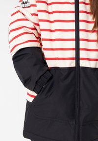 Rot-weiß gestreifte Jacke mit einem schwarzen unteren Abschnitt, ausgestattet mit elastischen Bündchen und einem vorderen Reißverschluss. Logo-Patch auf der oberen linken Seite.