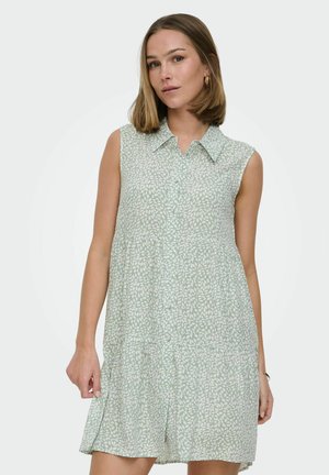 Ung kvinne iført ermeløs, lysegrønn kjole med hvitt bladmønster, står foran ensfarget lys bakgrunn.