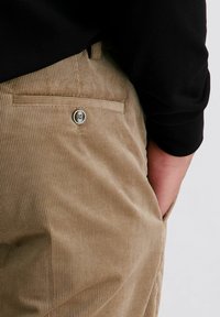 Beige corduroy broek met een achterzak met knoop; hand in de voortzak; zwart shirt met lange mouwen deels zichtbaar tegen een witte achtergrond.