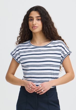 Tânără cu păr lung și creț poartă un top cu mâneci scurte alb cu dungi bleumarin și pantaloni bleumarin, cu mâinile încrucișate pe talie, arătând inele argintii.