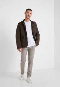 Casaco acolchoado castanho com fecho éclair, usado sobre uma t-shirt branca. Combinado com calças xadrez cinza claro e ténis brancos.