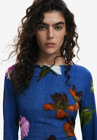 Bluse mit floralen Druck in denimähnlicher blauer Textur, mit lebhaften pinken, orangen und weißen Blumen sowie einem Bootsausschnitt.
