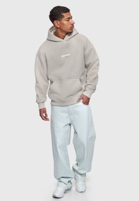 Joven mirando hacia la izquierda, vistiendo una sudadera con capucha beige con el texto "Dropsize", jeans anchos azul claro y zapatillas blancas sobre un fondo liso.