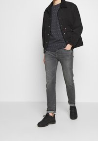 Černá džínová bunda s beránkovým límcem, tmavě modré tričko s vzorem, šedé džíny a černé tenisky, s volným střihem.