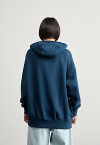 Persona con cabello oscuro y corto, vistiendo una sudadera azul oscura y jeans azules claros, de pie frente a un fondo liso de color gris claro.