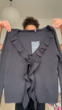 Άτομο κρατά μια μαύρη μπλούζα με μακριά μανίκια, με βολάν στο V-λαιμό και κορδελάκι μπροστά, δείχνοντας την ετικέτα της μάρκας με την ονομασία "ONLY".