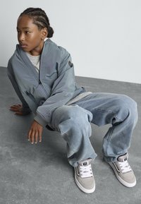 Hallide bomber-jakk, hall särk, laiad sinised teksad ja hallid Vans-sneakerid. Palmikutega juuksed, istub hallil pinnal neutraalse taustaga.