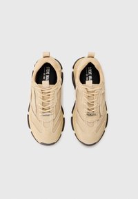 Zapatos deportivos beige con paneles de malla y ante, suelas y cordones negros contrastantes. Presenta una tira en el talón y un logo en la lengüeta.