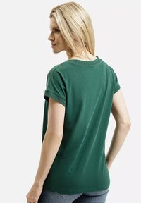 Blondynka w ciemnozielonym t-shircie z krótkim rękawem, widoczna od tyłu, w jasnoniebieskich dżinsach na jednolitym tle.