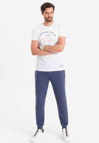 Bílý bavlněný tričko s modrým a oranžovým grafickým textem, spojené s námořnickými tepláky s ribbovanou texturou a bočními kapsami. Bílé tenisky.