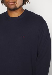 Navy knitted sweater with a crew neckline. Displays a small red, white, and blue logo on the left chest. Smooth texture, medium weight fabric.
