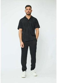 Une chemise noire à manches courtes et un pantalon assorti présentent un motif en vague texturé, avec une coupe décontractée, des boutons à l'avant et des baskets blanches.