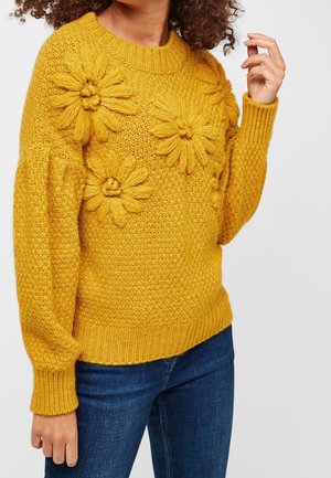 Pull en tricot jaune moutarde avec de grands motifs floraux texturés, porté avec un jean bleu, visible du milieu du torse jusqu'au cou et aux bras.