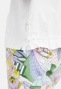 Weißes, leichtes Oberteil mit gerafftem Saum und Logo-Detail, kombiniert mit bunten, gemusterten Leggings mit floralen und abstrakten Designs.