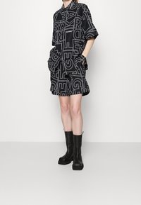 Osoba v černé oversized košili a šortkách s bílými grafickými písmeny, doplněná černými masivními kozačkami po lýtka, stojící na bílém pozadí.