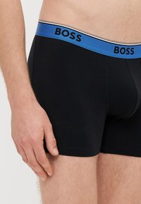 Μαύρα ανδρικά εσώρουχα τύπου boxer με μπλε και χρυσή ελαστική μέση που φέρει το λογότυπο "BOSS". Λείο ύφασμα με εφαρμοστό σχέδιο.