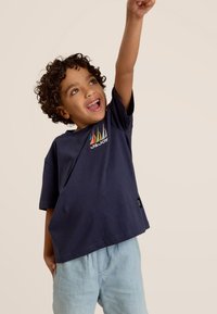 Marineblauwe katoenen t-shirt met een kleurrijke zeilbootprint en korte mouwen, gecombineerd met lichtblauwe shorts, tegen een neutrale achtergrond.