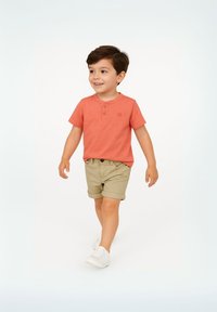Korallfärgad t-shirt med kort ärm och knappad hals, i kombination med kaki shorts. Vita sneakers fulländar outfiten. Barnet går på en neutral bakgrund.