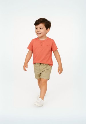 Chemise corail à manches courtes avec un col boutonné, associée à un short kaki. Des baskets blanches complètent la tenue. L'enfant marche sur un fond uni.