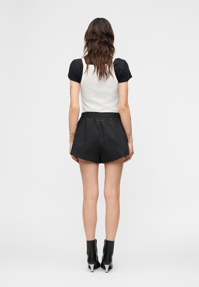 Γυναίκα που στέκεται με την πλάτη προς την κάμερα, φοράει μαύρο κοντομάνικο τοπ, λευκό αμάνικο γιλέκο, μαύρες σορτσάκια και μαύρα μποτάκια με τακούνι.