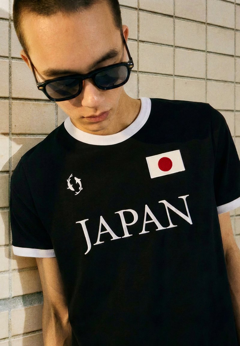 Young man wearing sunglasses and a black T-shirt with white trim, featuring the word "JAPAN" and a Japanese flag patch.