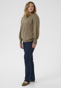 Vrouw die staat in een olijfgroene trui met pareldetails, donkere spijkerbroek en bruine flats, haar hoofd iets naar links gekanteld.