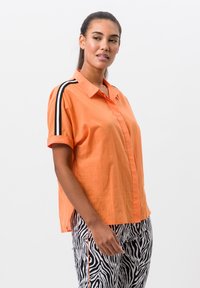 Korte mouwen oranje shirt met een knoopsluiting aan de voorkant, met zwart-witte gestreepte accenten op de schouders. De stof lijkt lichtgewicht en gestructureerd.