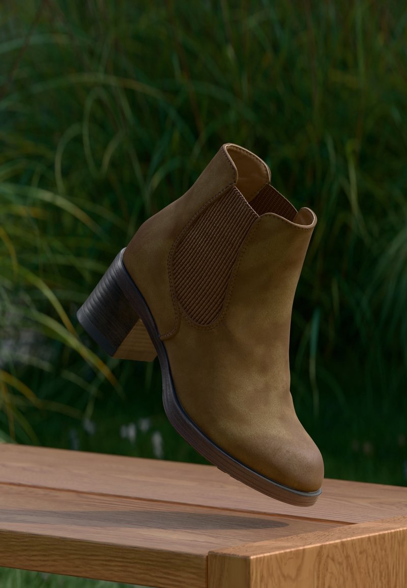 Bottine en daim marron avec panneaux latéraux élastiques et talon superposé, suspendue au-dessus d'une surface en bois, fond de feuillage vert.