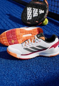 Witte sportieve schoenen met oranje en bordeaux accenten, met een ademende mesh bovenkant, een gestructureerde zool en een opvallend drie-strepen ontwerp.