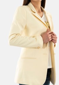 Blazer de color crema con un corte entallado, solapas con muesca y acentos de botones dorados, combinado con una camiseta gráfica blanca por debajo.